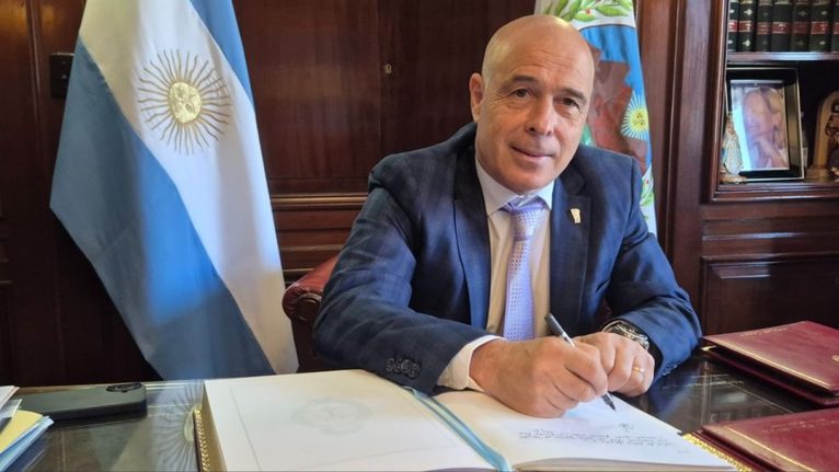Funcionario firma documentos en un despacho junto a la bandera argentina