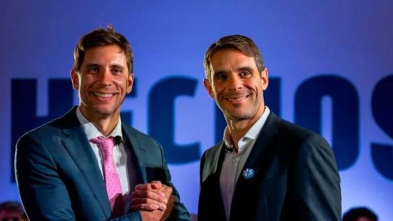 Dos dirigentes de Hechos posan frente a un fondo azul con el logo del espacio